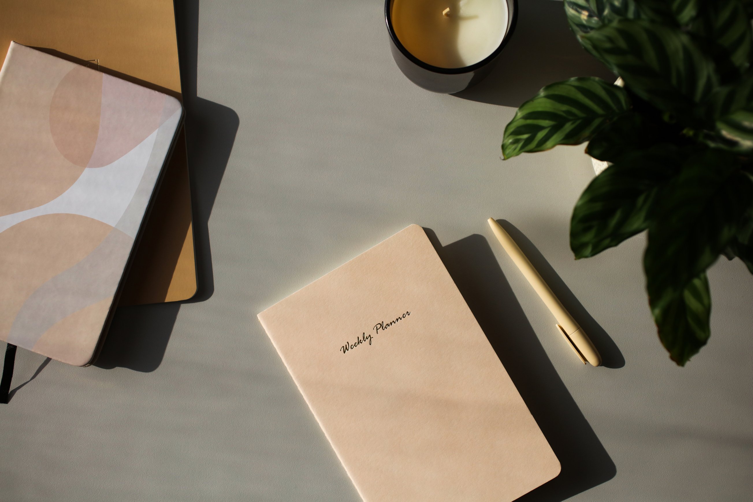 Aesthetic Feminine Workspace with Weekly Planner, Notebooks, Pen, Candle, Green Plant on Table.